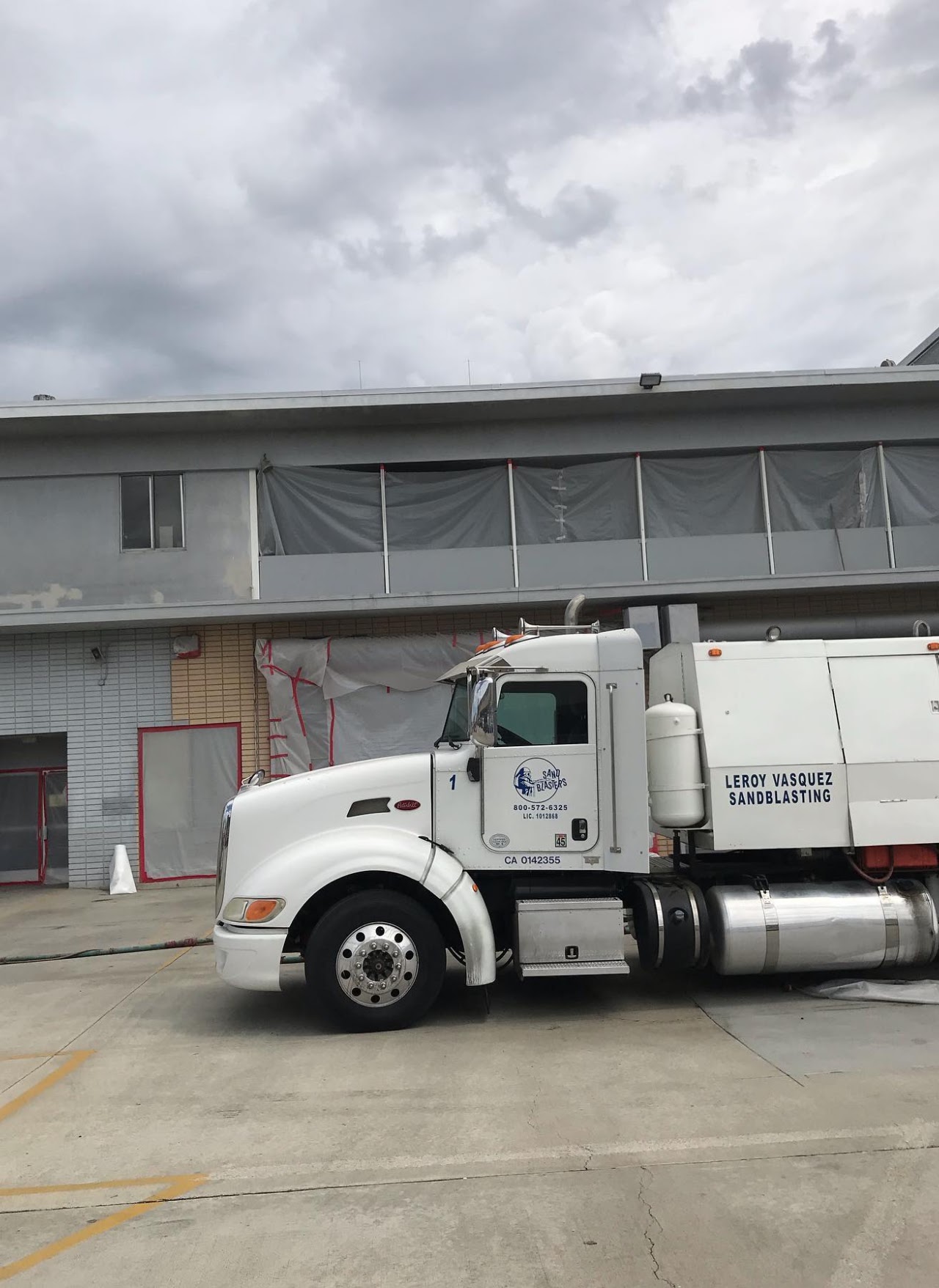 A white Leroy Vasquez Sandblasting semi truck parked outside a warehouse