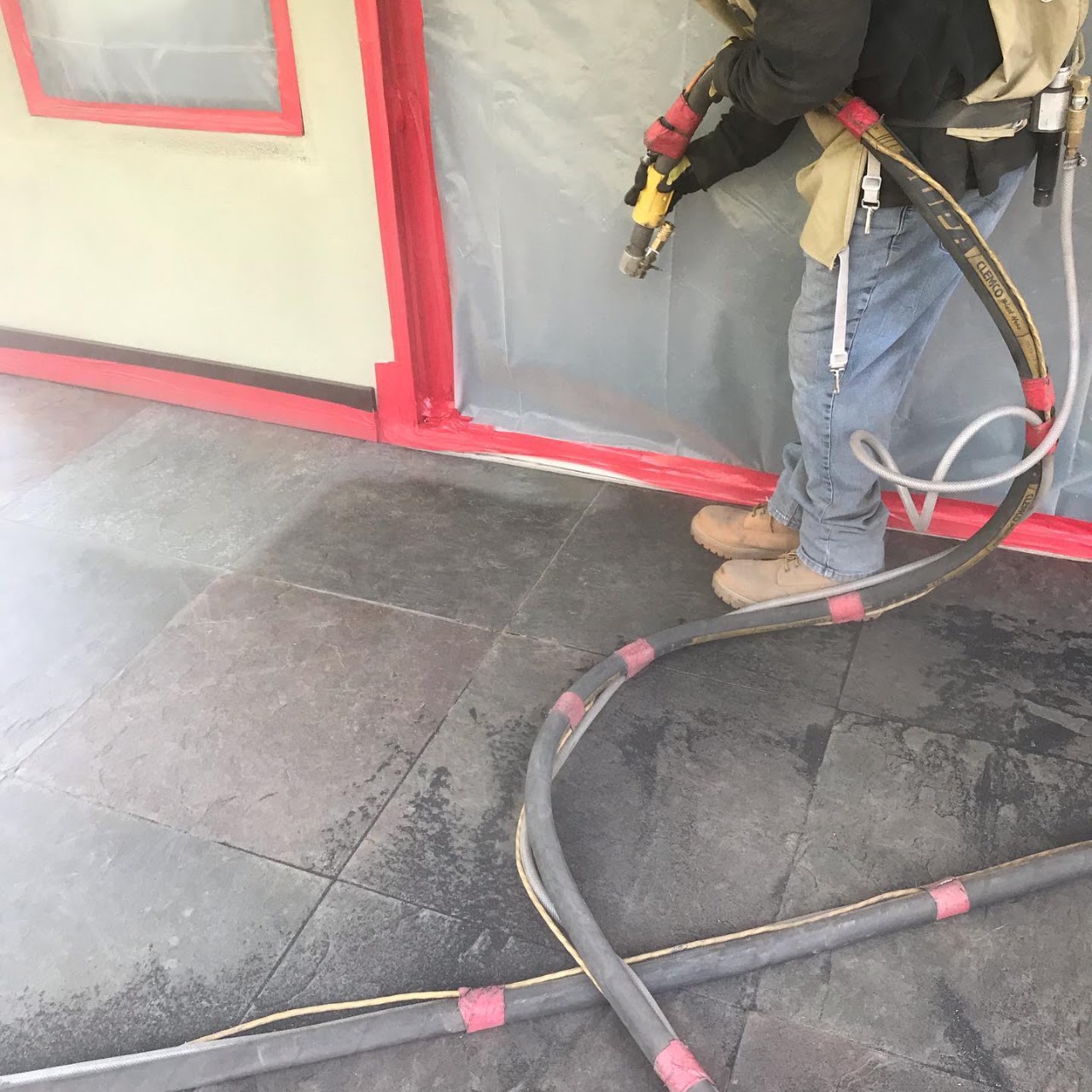 A sandblasting operator in protective gear working on a tiled floor inside a plastic-sheeted containment area